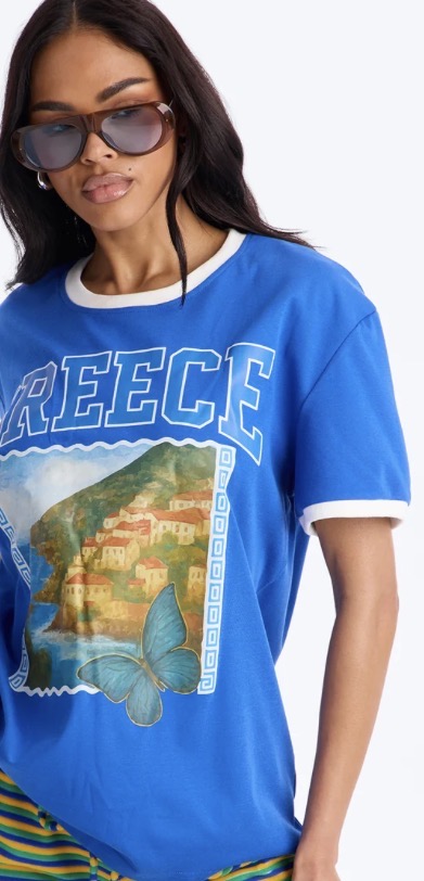 Model wearing a royal blue ringer tee with a painted Greek coastal village and butterfly graphic