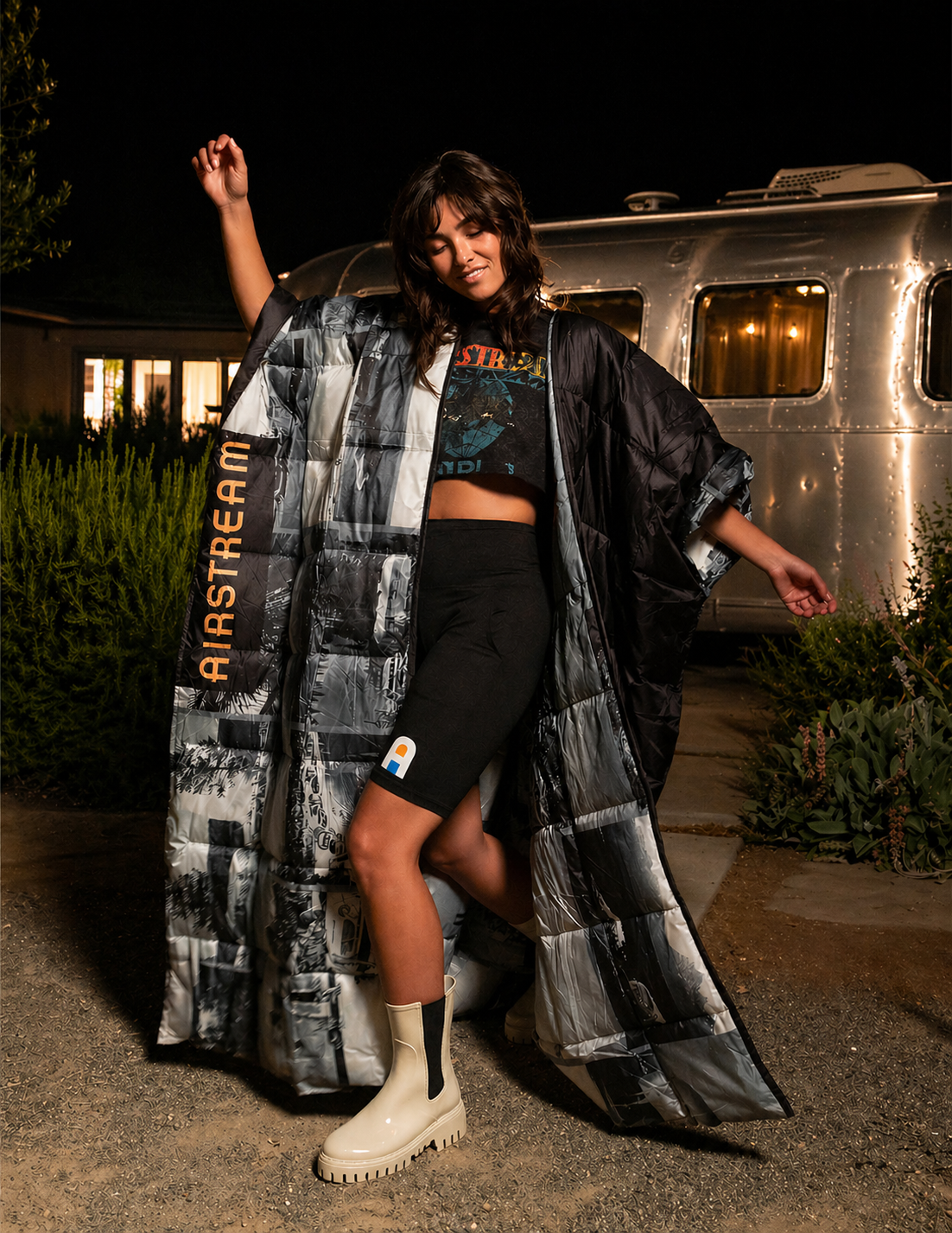 Model wearing an oversized Airstream puffer poncho printed with black-and-white trailer imagery at night
