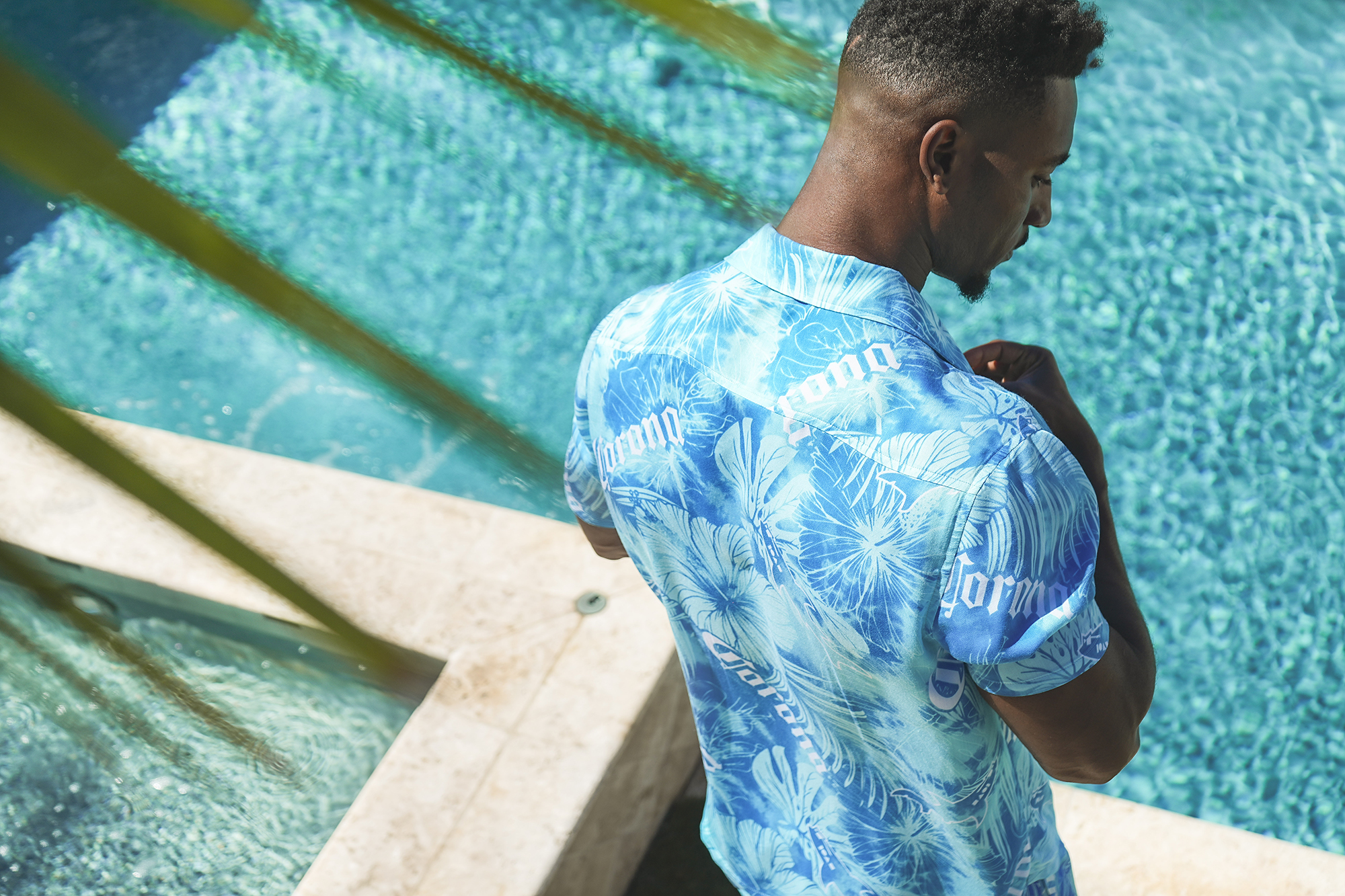 Model wearing a blue Corona and Maui and Sons Palm Fusion Hawaiian shirt beside a swimming pool